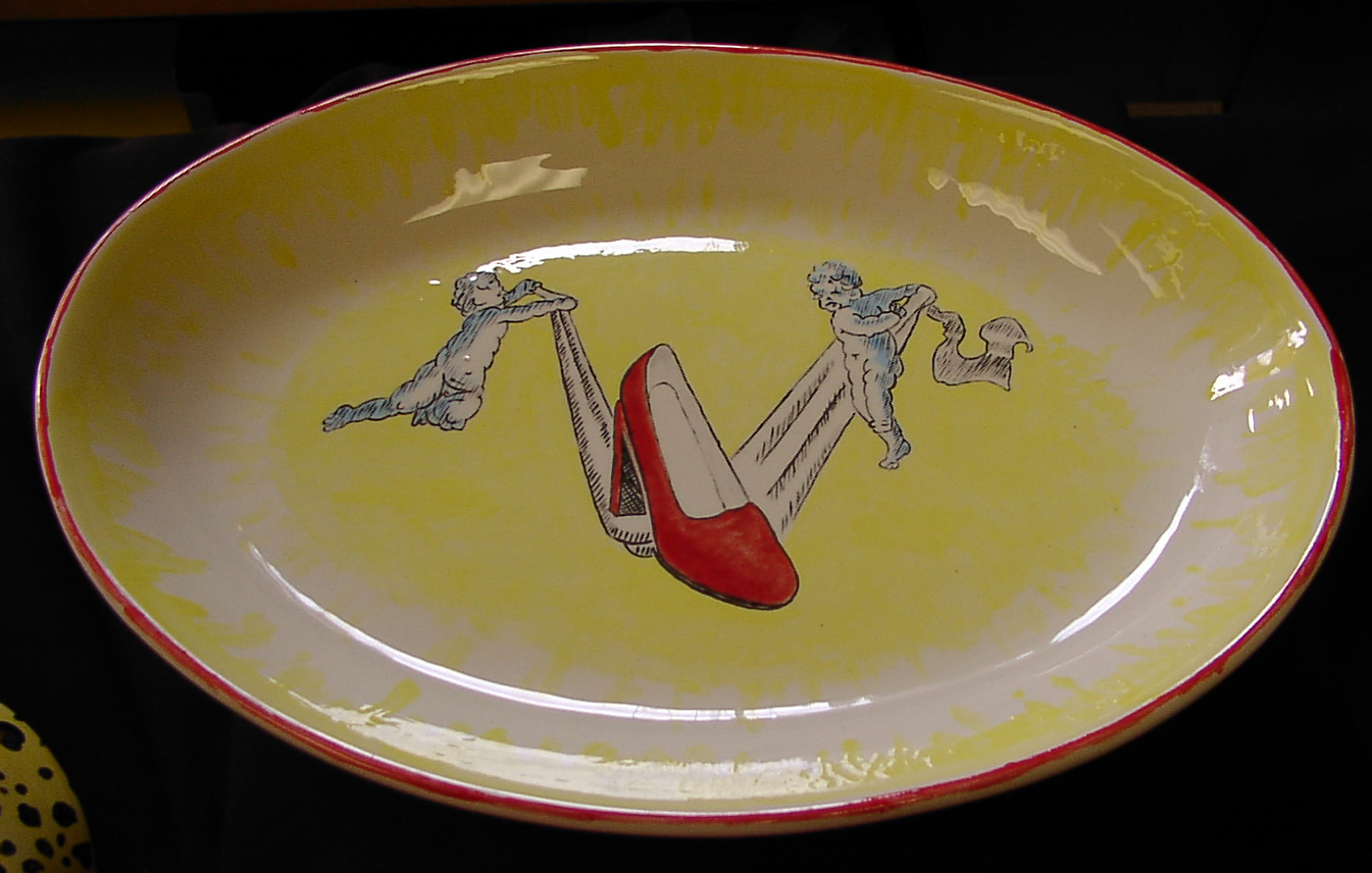 An oval platter decorated with two putti supporting an enormous red, high-heeled shoe.